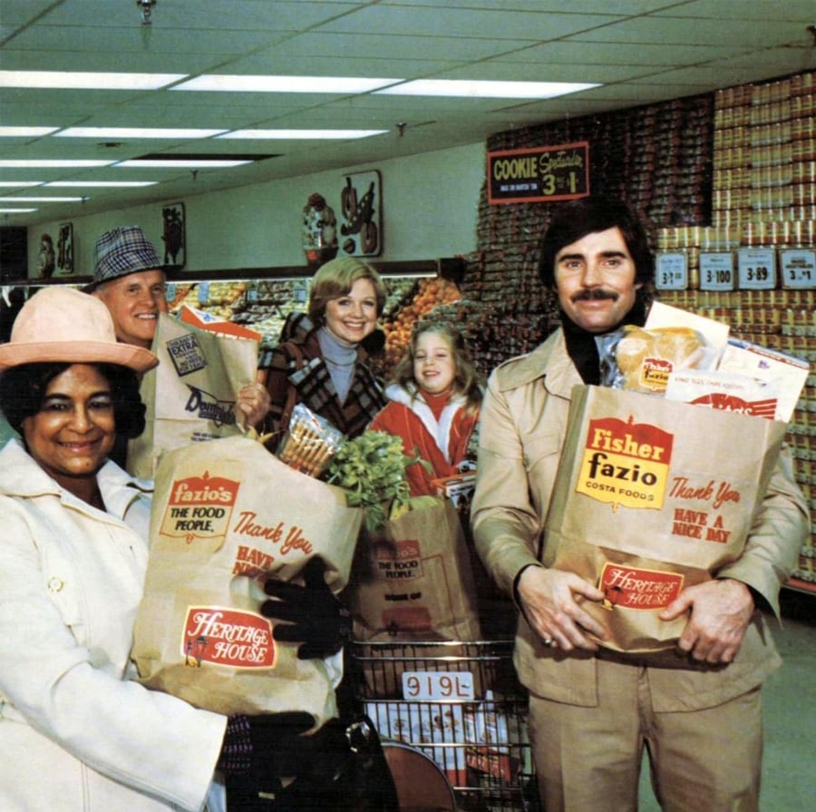 A group of smiling people stand in a grocery store aisle, holding paper bags filled with groceries. Shelves stocked with canned goods and a cookie sign are visible in the background.