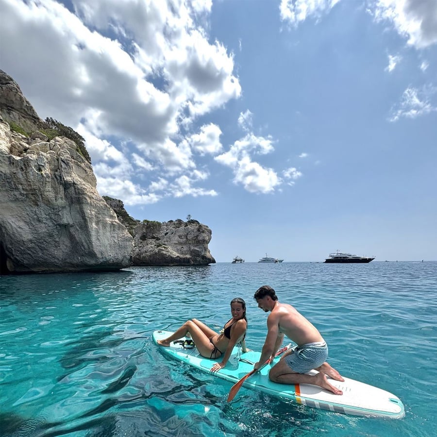 A woman sits on a paddleboard while a man kneels and paddles beside her on clear blue water near rocky cliffs, with several boats in the distance under a partly cloudy sky.