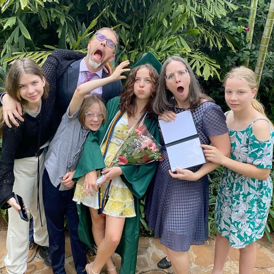 A group of six people, including a young graduate in a green gown holding flowers, stand together outdoors making playful and surprised faces, surrounded by greenery.