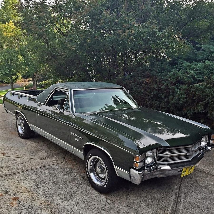 A vintage green Chevrolet El Camino parked on a driveway, surrounded by trees and bushes on a sunny day.