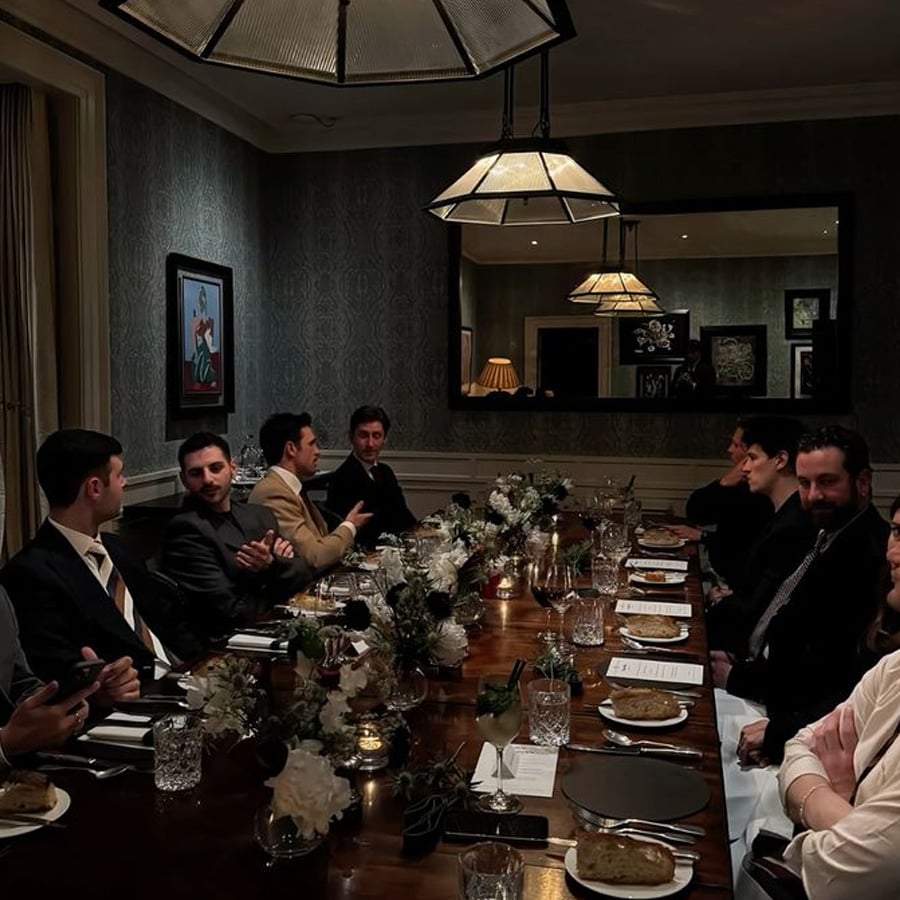 A group of people dressed formally sit around a long dining table set with plates, glasses, flowers, and candles in a dimly lit, elegant room with dark wallpaper and framed art.