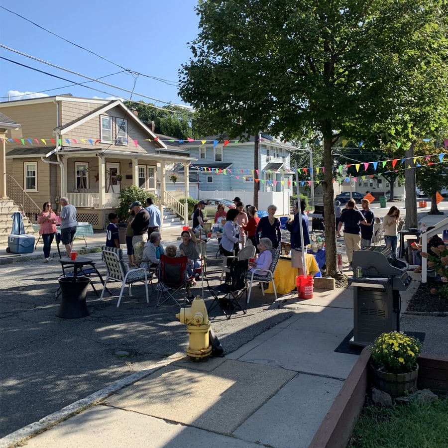 People gather on a residential street for a block party, sitting and standing near tables with food and drinks. Colorful pennant banners hang overhead, and houses line the background on a sunny day.