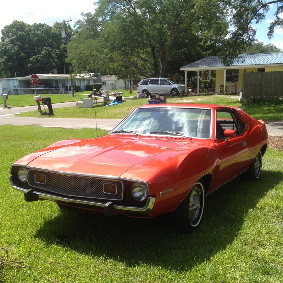 A bright red vintage car is parked on a grassy lawn in front of a house with trees and a stop sign visible in the background on a sunny day.