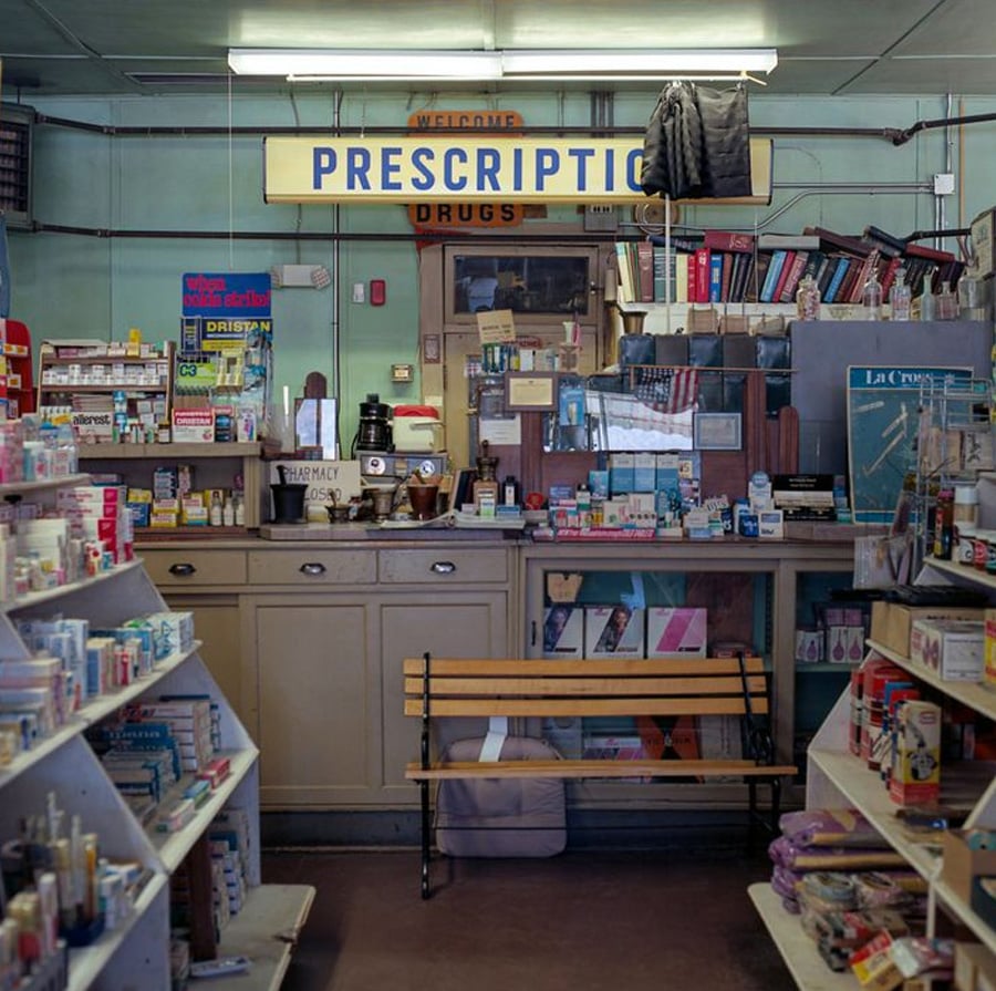 A vintage pharmacy interior with shelves stocked with medicines and products, a "PRESCRIPTION DRUGS" sign overhead, a wooden bench in front of the counter, and various items and books behind the counter.