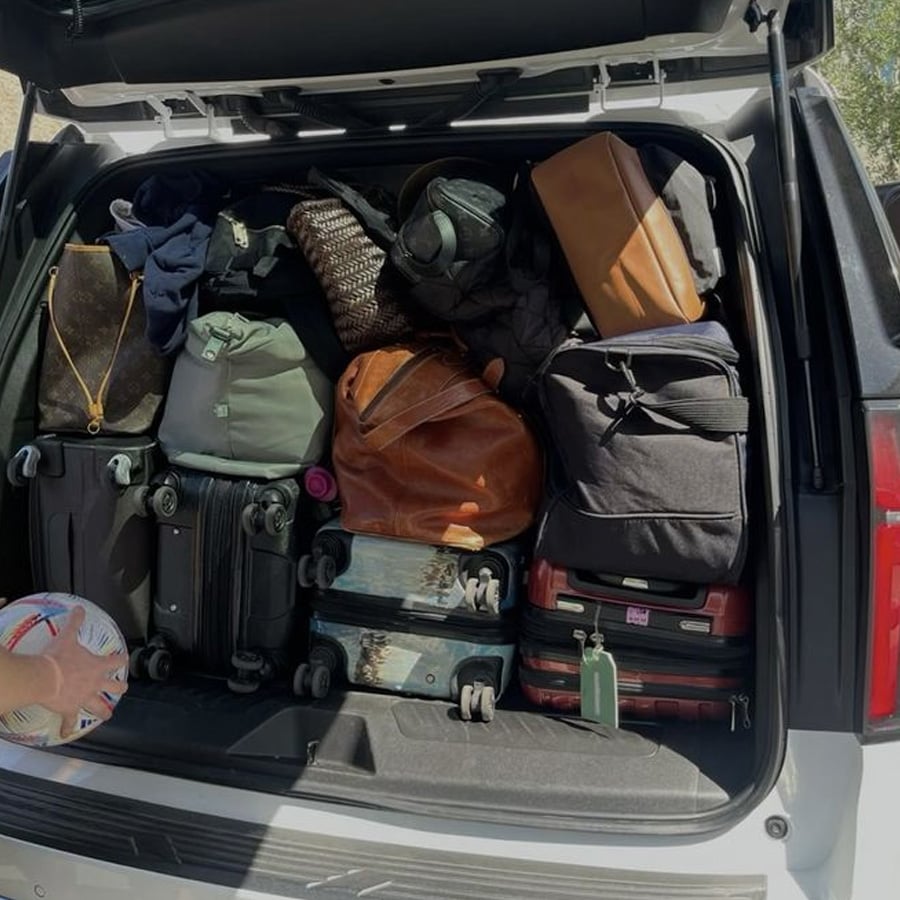 An SUV trunk packed tightly with various suitcases, bags, and backpacks, some with wheels. A hand holds a frisbee to the left edge of the open trunk, under bright sunlight.