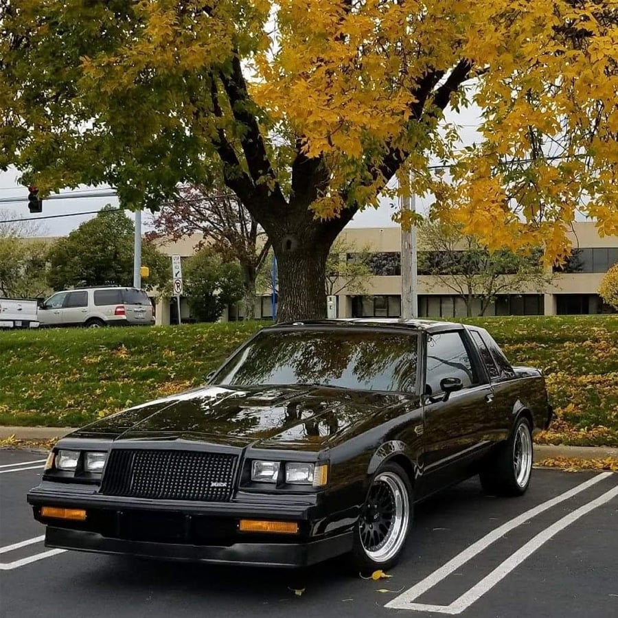 A vintage black car is parked in a lot under a tree with yellow autumn leaves, with office buildings and other vehicles visible in the background.