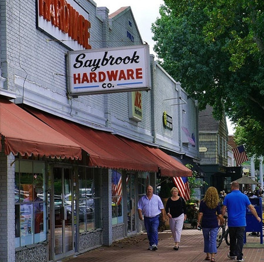 People walk along a sidewalk in front of Saybrook Hardware Co. and other shops on a tree-lined street with red awnings and store signs visible on the buildings.