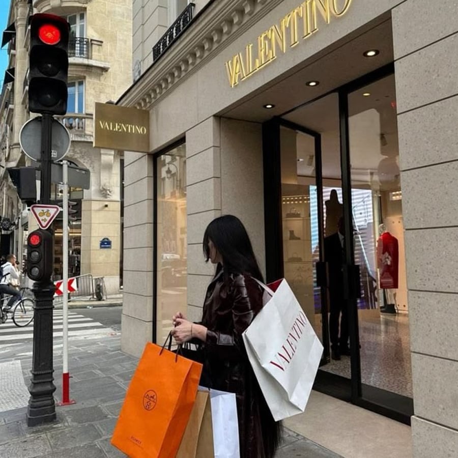 A woman with long dark hair, wearing a dark coat, stands outside a Valentino store holding multiple shopping bags. The street corner has a traffic light and classic Parisian architecture in the background.