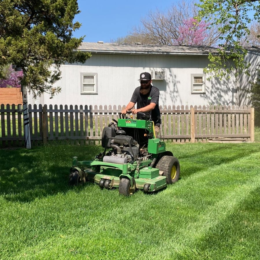 A person wearing a black shirt and hat is mowing a green lawn with a large green lawn mower in a sunny backyard, with a wooden fence and a white shed in the background.