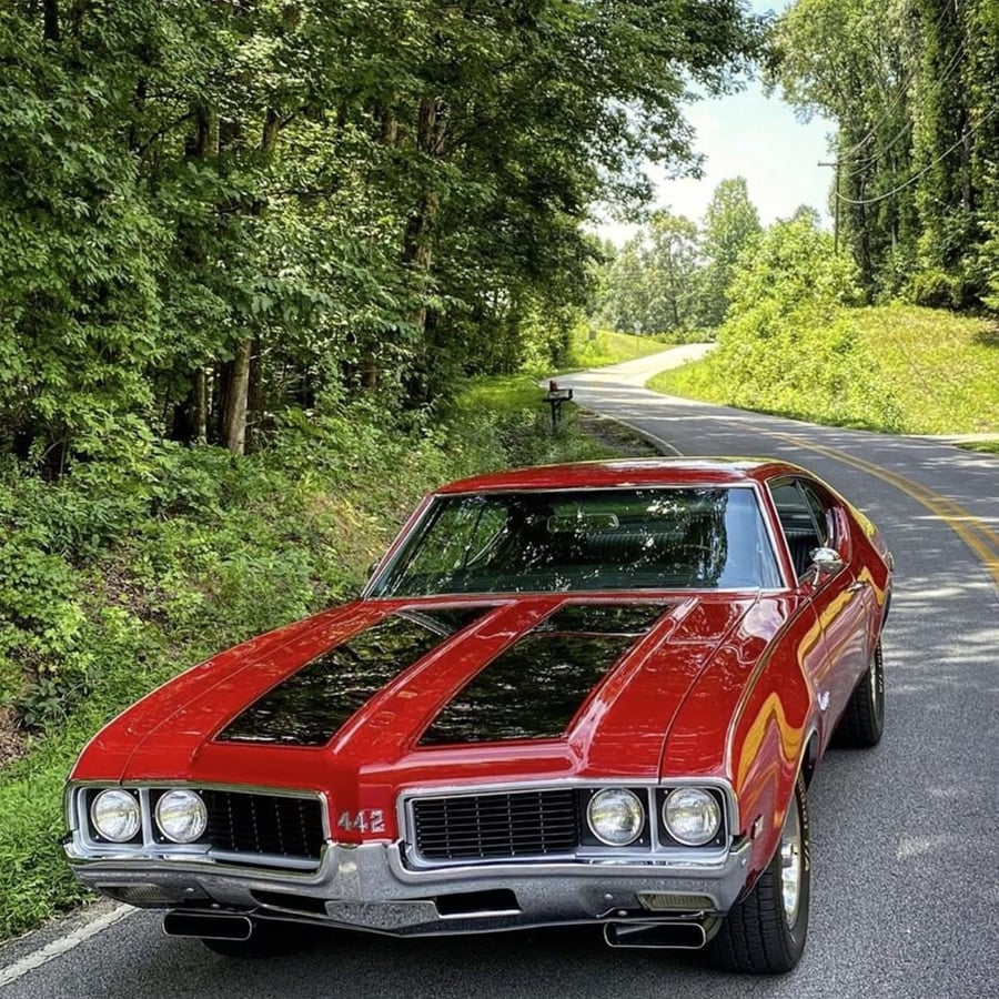 A classic red muscle car with black racing stripes is parked on the side of a winding road surrounded by green trees and foliage on a sunny day.