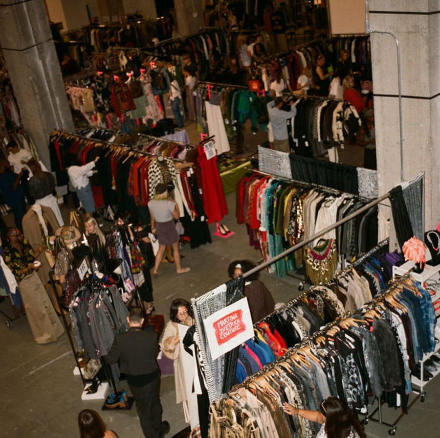A large indoor market with rows of clothing racks filled with colorful clothes, and many people browsing and shopping in the busy space. Signs and concrete pillars are visible throughout the area.