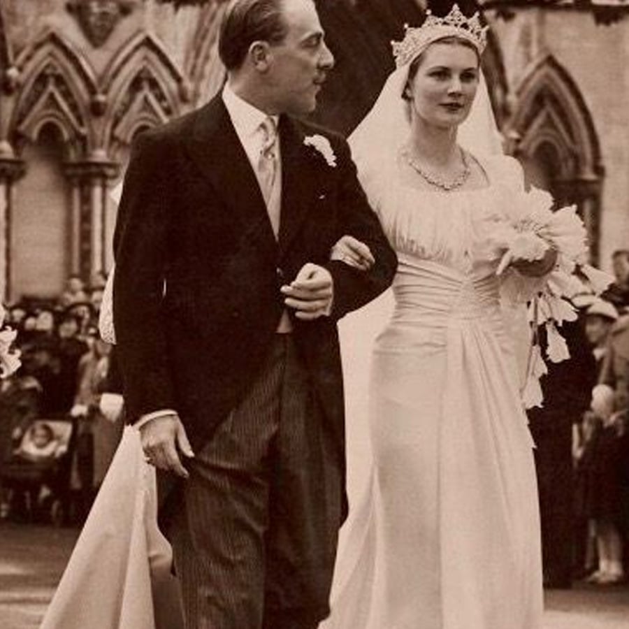 A bride in a white gown and tiara walks arm in arm with a man in formal attire, in front of a crowd and a gothic-style building, during a wedding ceremony.