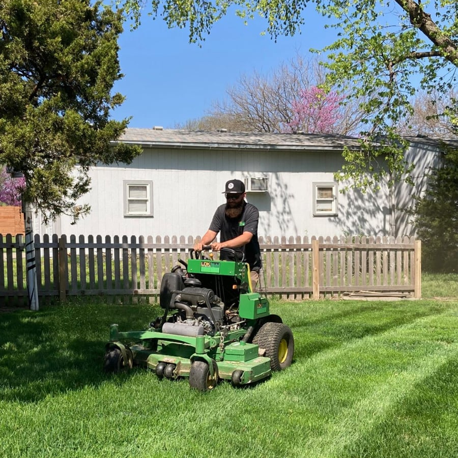 A person wearing a cap and sunglasses is mowing a green lawn with a large green lawn mower in a sunny backyard, with a wooden fence, trees, and a white building in the background.
