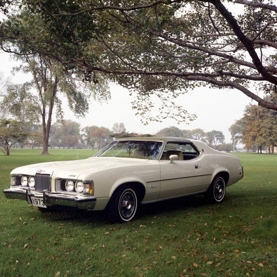 A vintage white 1971 Mercury Cougar coupe is parked on green grass under large, leafy trees in a park. The car has a vinyl roof, chrome accents, and a classic design, with a license plate reading "1971.