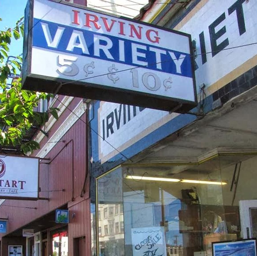 A vintage storefront sign reads “Irving Variety 5¢ & 10¢” above a shop entrance. Reflections and neighboring storefronts are visible along the street on a sunny day.