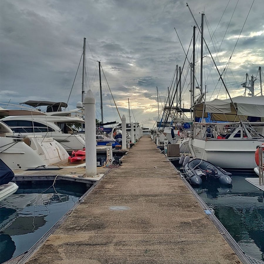 A concrete dock extends into the distance, lined with docked sailboats and yachts on both sides under a cloudy sky, with calm water reflecting the boats and sky.