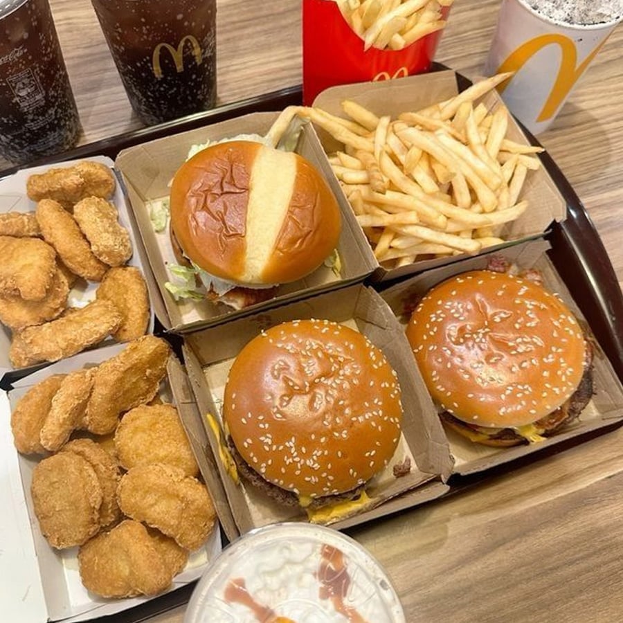 A tray filled with McDonald’s food: three burgers, a box of chicken nuggets, fries, two cups of soda, and a cup with a dome lid, all placed on a wooden table.
