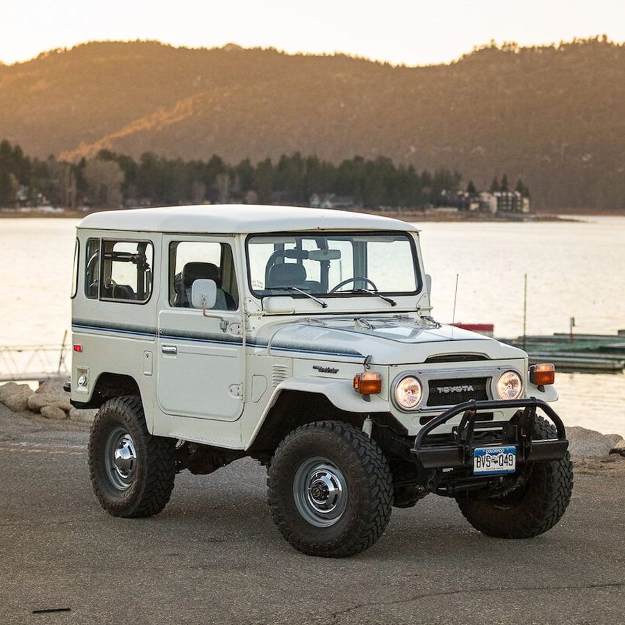 A white classic Toyota Land Cruiser is parked near a lake at sunset, with mountains and trees in the background. The vehicle has large tires and a vintage design.