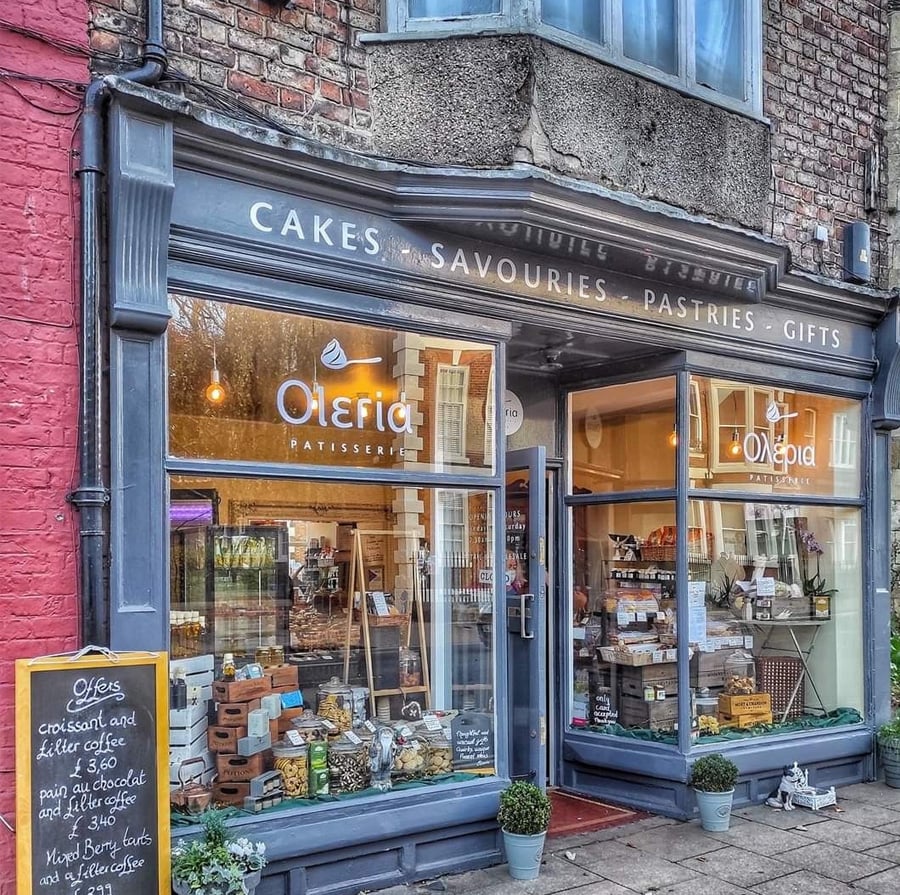 A charming bakery storefront with large windows displays cakes, pastries, and gifts. A sign outside lists menu specials, and the shop is named "Olefia Patisserie." The exterior has grey trim and a welcoming atmosphere.