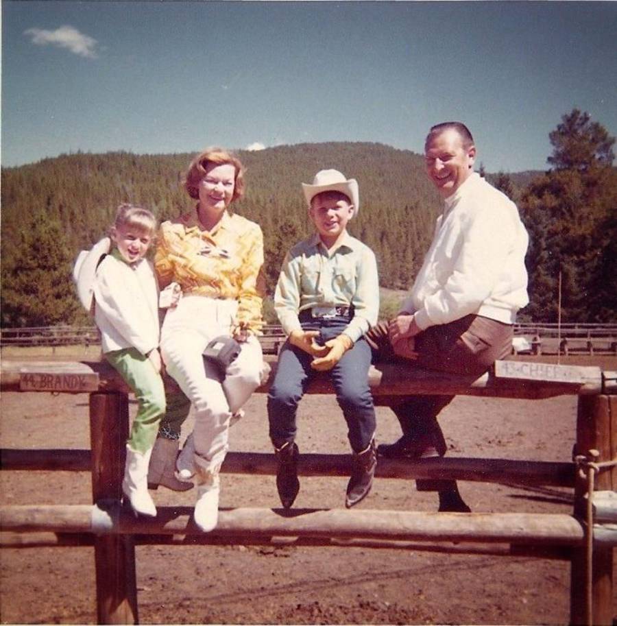 A family of four sits on a wooden fence at a ranch, dressed in colorful western-style clothing. Trees and hills fill the background under a clear sky. Everyone is smiling and enjoying the sunny day.