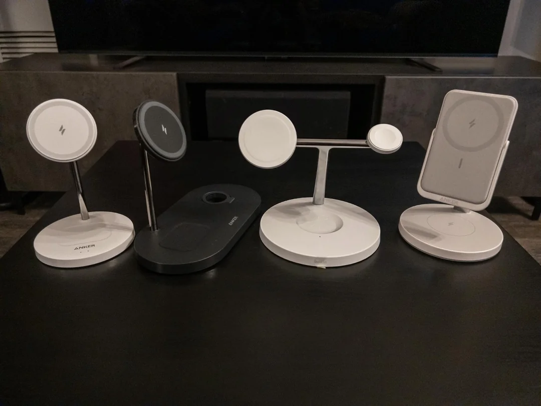 Four wireless charging stands with round charging surfaces are displayed side by side on a black table in front of a TV stand. The chargers are in different colors and designs, mostly white and black.