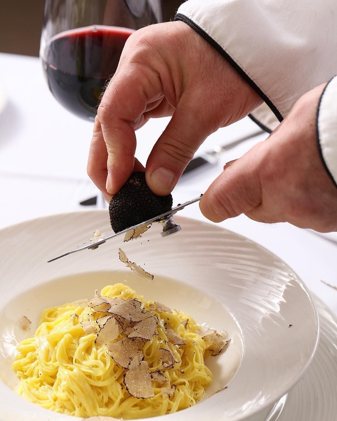 A chef shaves black truffles over a plate of fresh pasta, with a glass of red wine in the background on a white tablecloth.