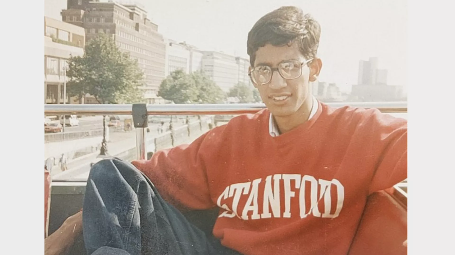 A young man with glasses wearing a red Stanford sweatshirt sits relaxed on an outdoor bench with buildings and trees in the background.