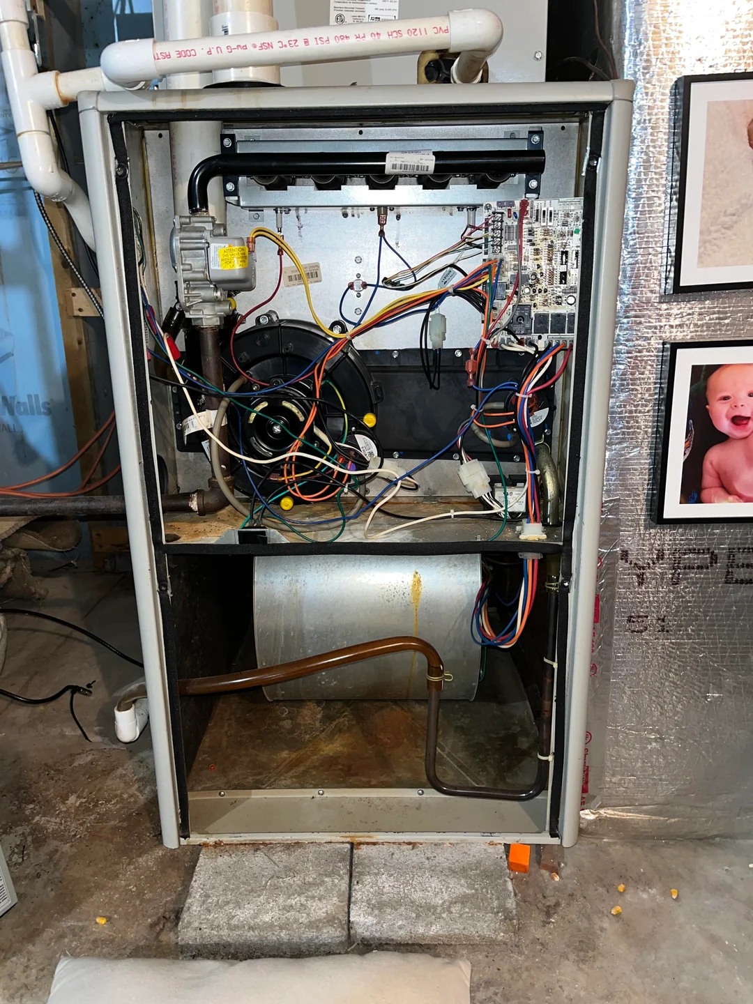 A furnace with its front panel removed, revealing internal wiring, circuit boards, and components. The unit sits on concrete blocks in a basement with exposed walls and photos on the right side.