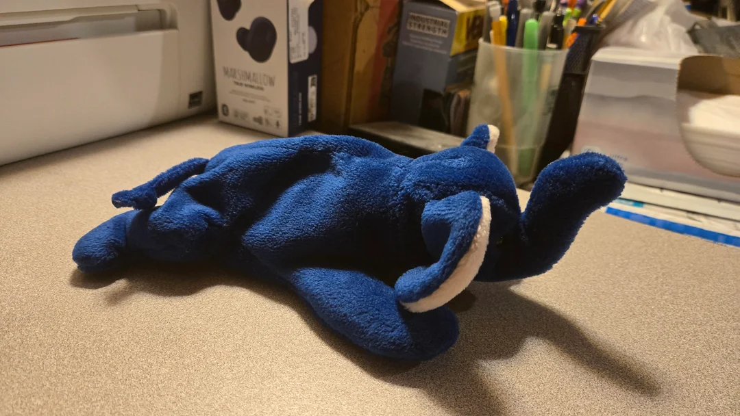 A blue plush elephant toy lies on its stomach on a desk, with its trunk raised. In the background are office supplies, books, and assorted desk items.