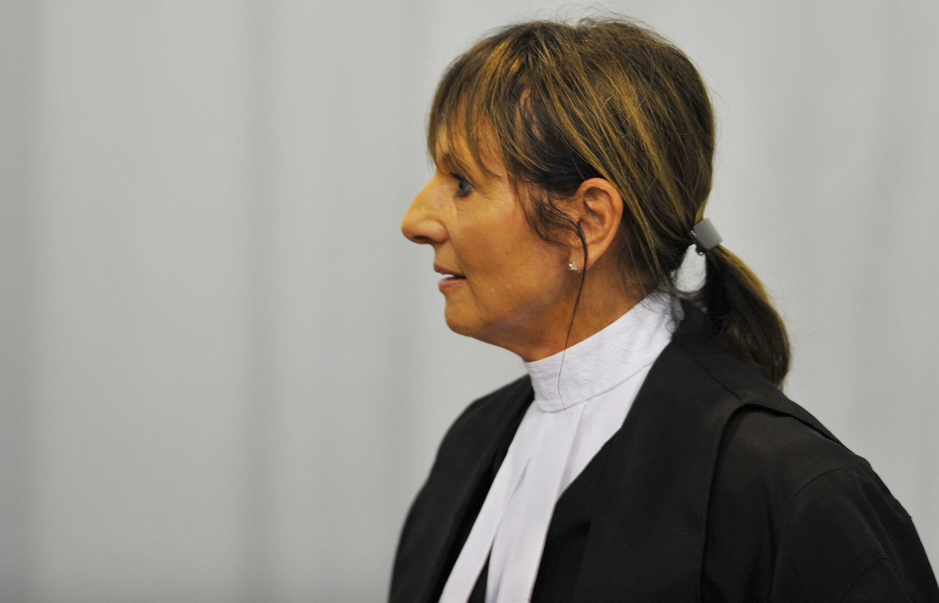 A woman wearing a black legal robe and white collar stands in profile, looking to the left. Her brown hair is tied back in a ponytail with a grey hair tie. The background is plain and out of focus.