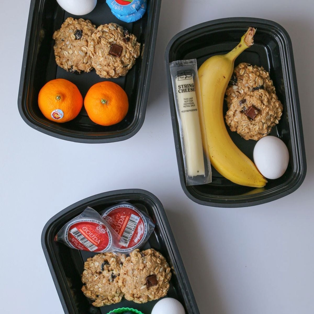 Three black meal prep containers, each holding two oatmeal cookies, a hard-boiled egg, and cheese. Two containers have mandarins; one has a banana and string cheese. All have individual cheese portions.
