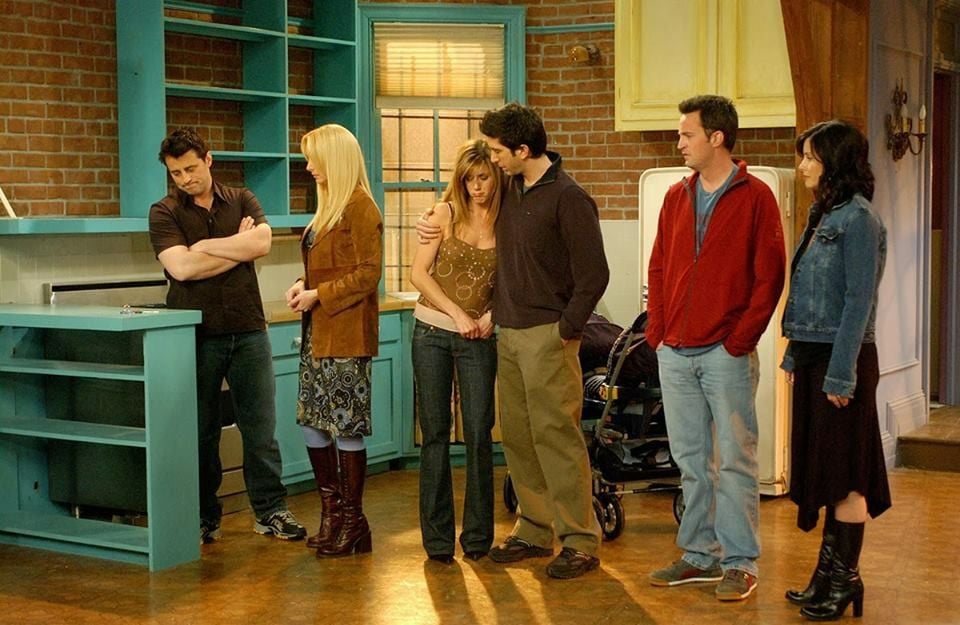Six people stand in a kitchen with teal cabinets, some hugging and looking emotional, while others stand nearby. The setting has brick walls and a window, giving a homey, apartment feel.