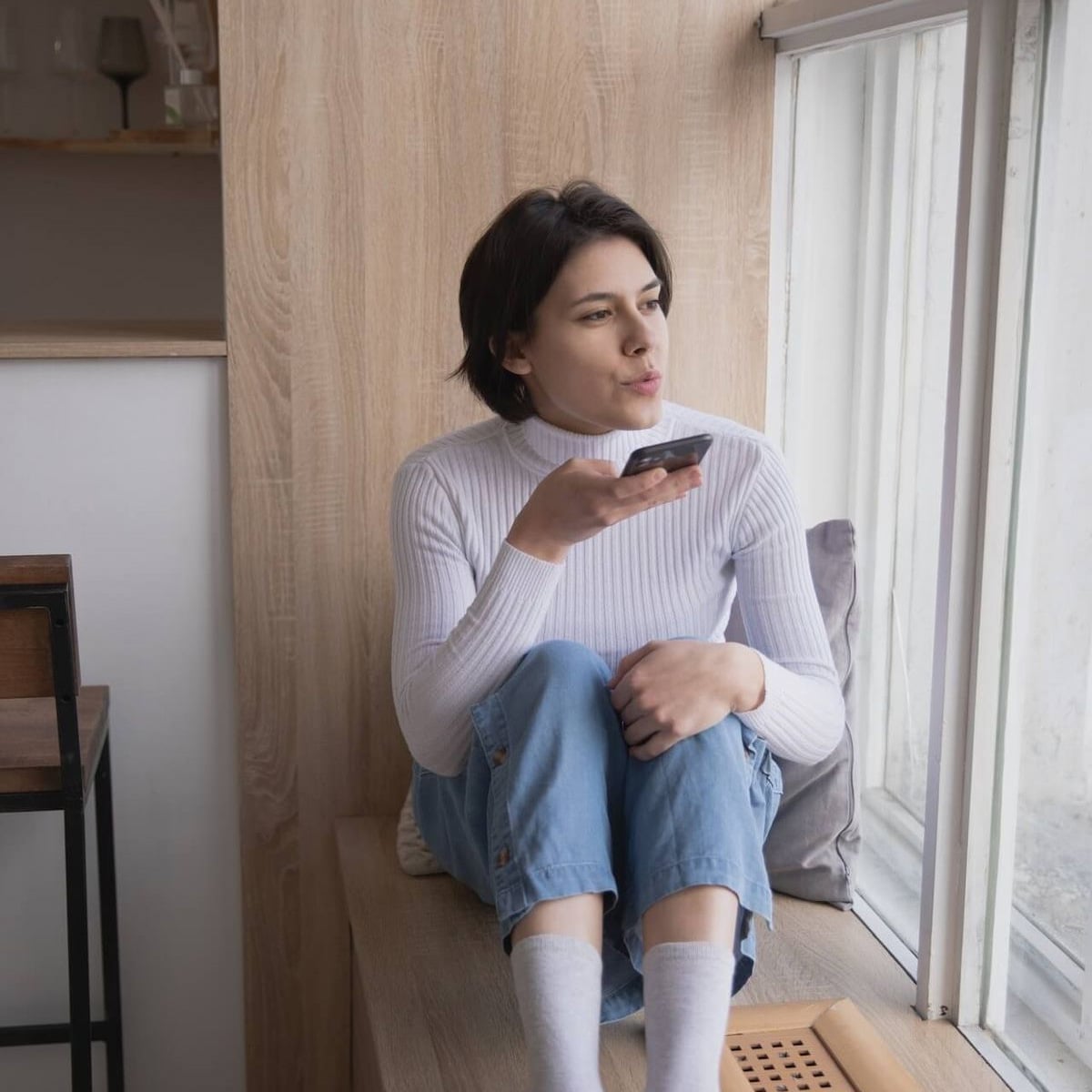 A person in a white turtleneck and jeans sits by a window, holding a phone and speaking into it, appearing relaxed and thoughtful.