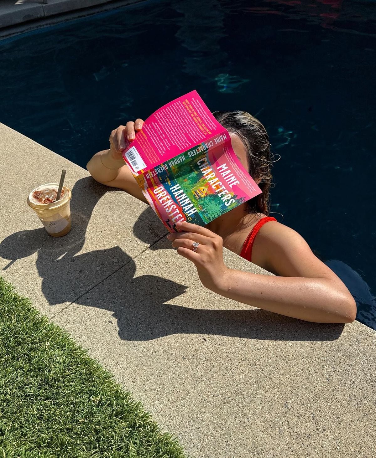 A person in a red swimsuit relaxes in a swimming pool, leaning on the edge while reading a colorful book. An iced coffee with a spoon rests on the poolside near the grass.
