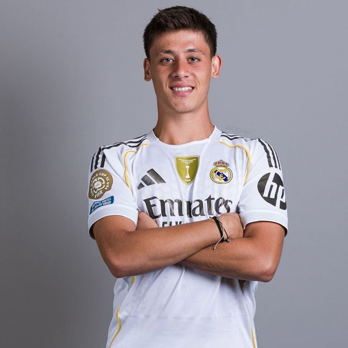 A young man wearing a Real Madrid soccer jersey stands with arms crossed, smiling at the camera against a plain gray background.