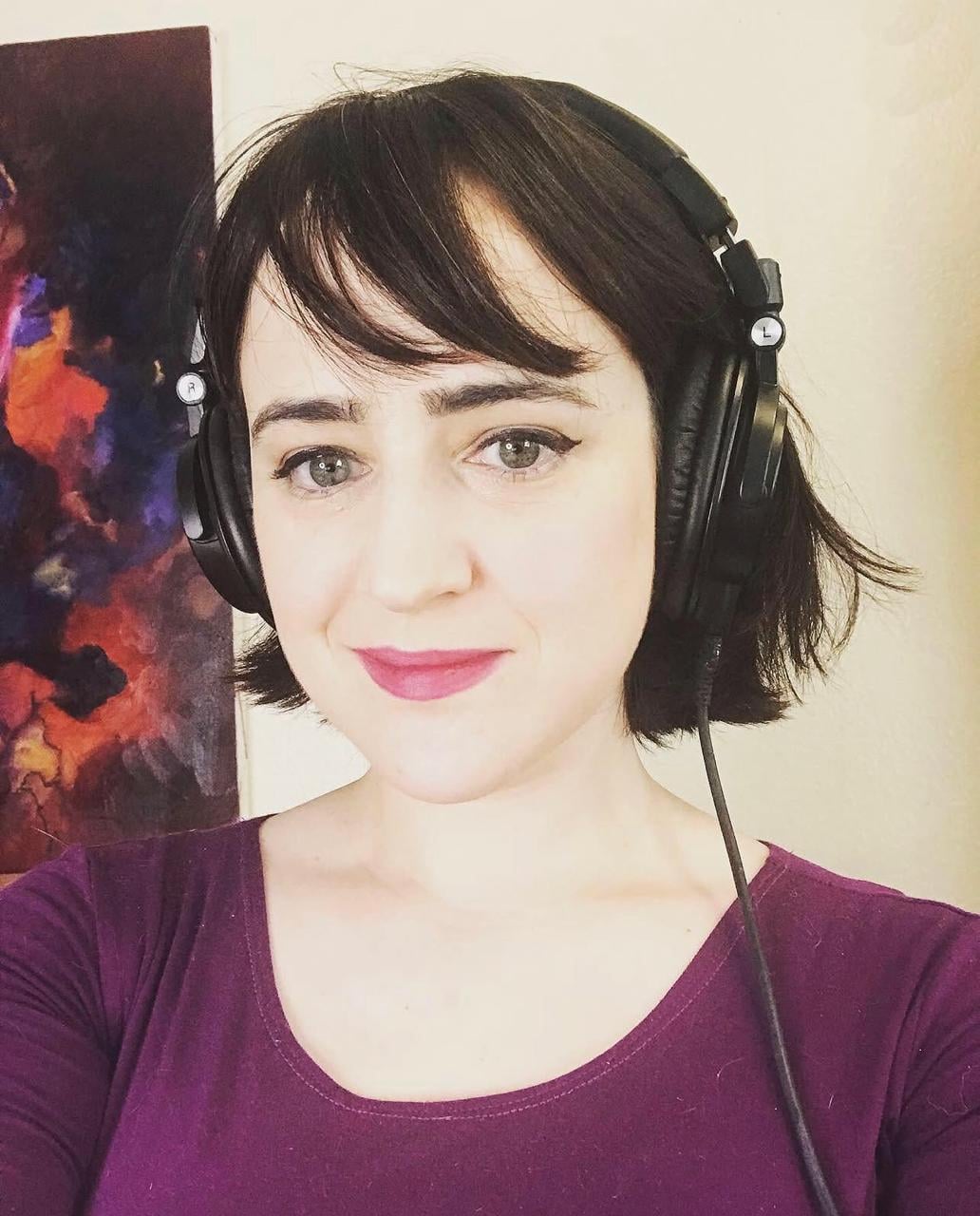A woman with short dark hair wearing large black headphones and a purple top smiles at the camera. There is a colorful abstract painting in the background.