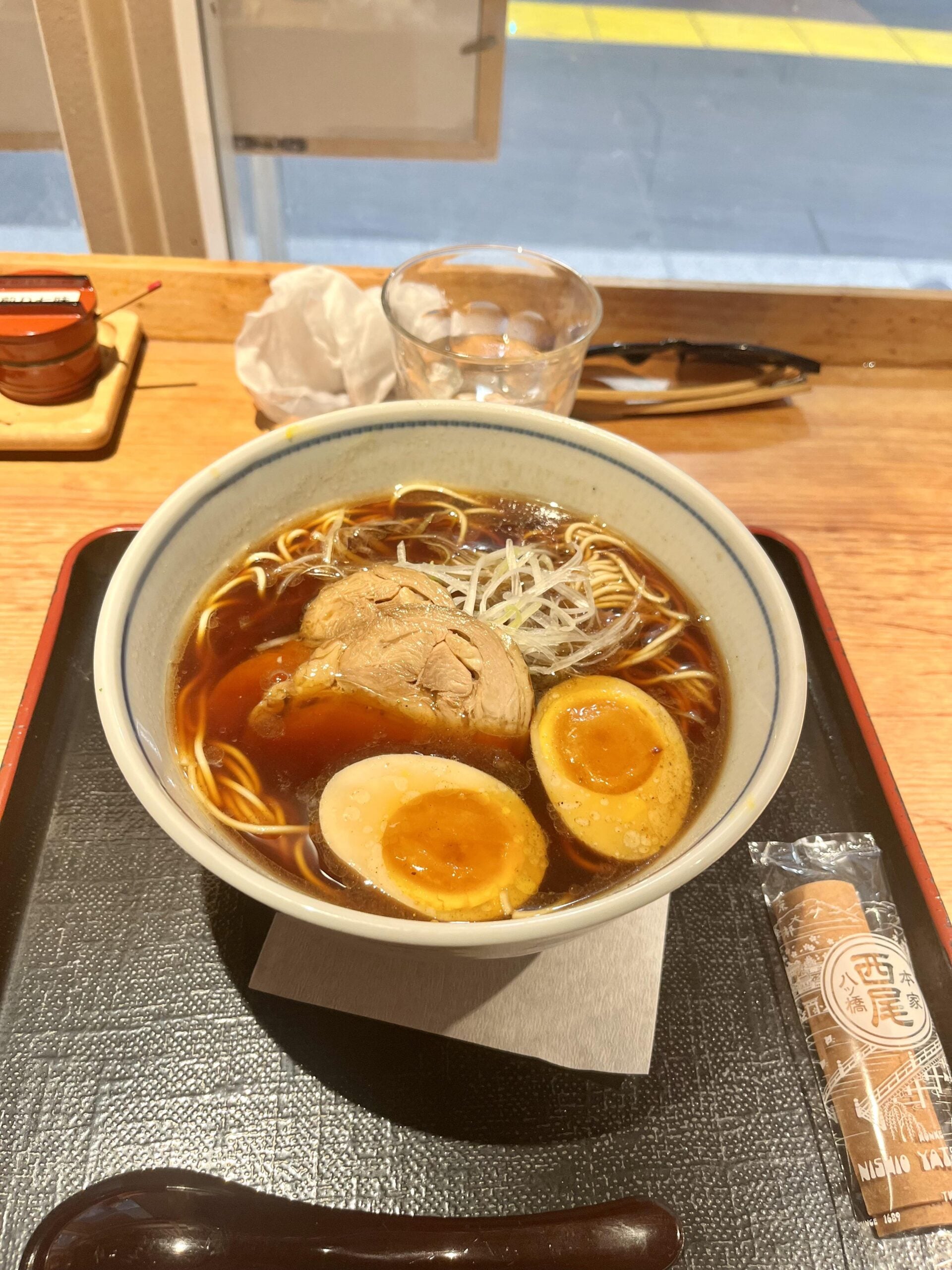 A bowl of ramen with sliced pork, two halved soft-boiled eggs, noodles, and green onions in broth sits on a tray with a napkin, chopsticks, spoon, a glass of water, and a wrapped snack on a wooden table by a window.