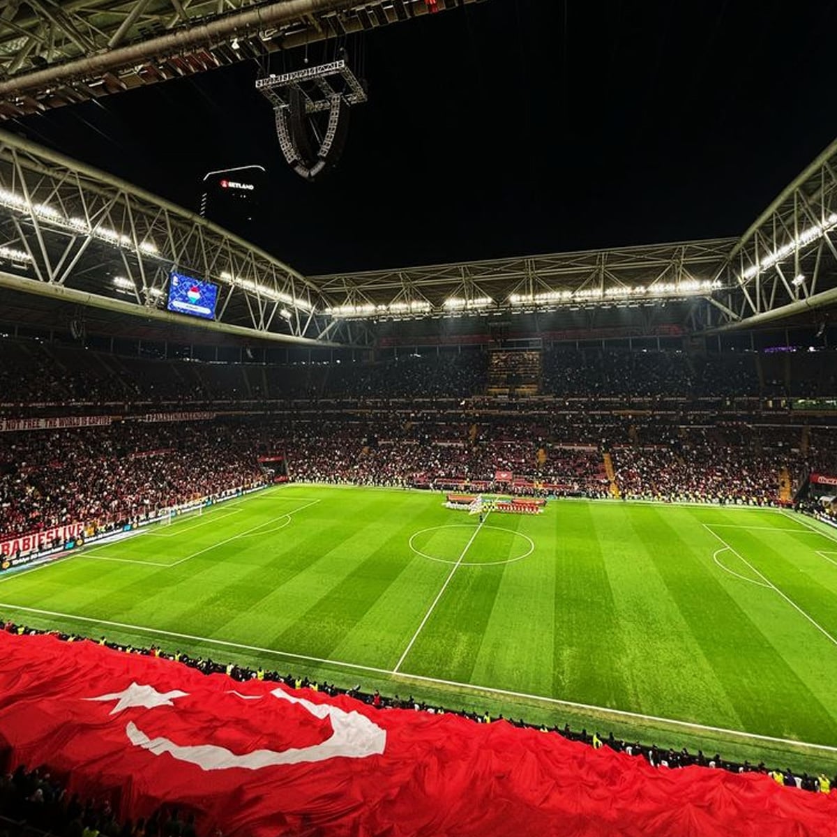 A large soccer stadium at night, filled with cheering fans. The well-lit field is ready for a match, and a huge red Turkish flag with a white crescent and star is displayed in the stands.