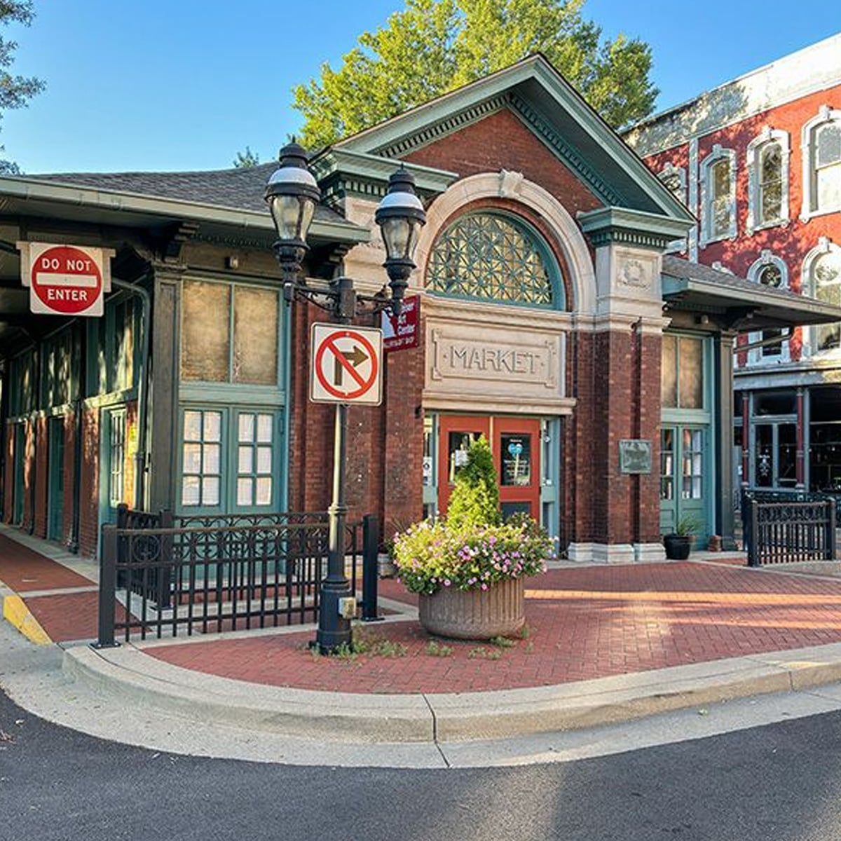 Historic brick market building with large arched window, lamp posts, and no left turn and do not enter signs. There’s a planter with greenery on red brick pavement and trees in the background.