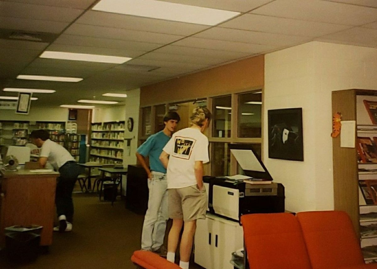 Three people are in a library. Two stand by a photocopier, with one lifting the lid, while the third person is at a counter in the background. Bookshelves and library furniture are visible throughout the room.