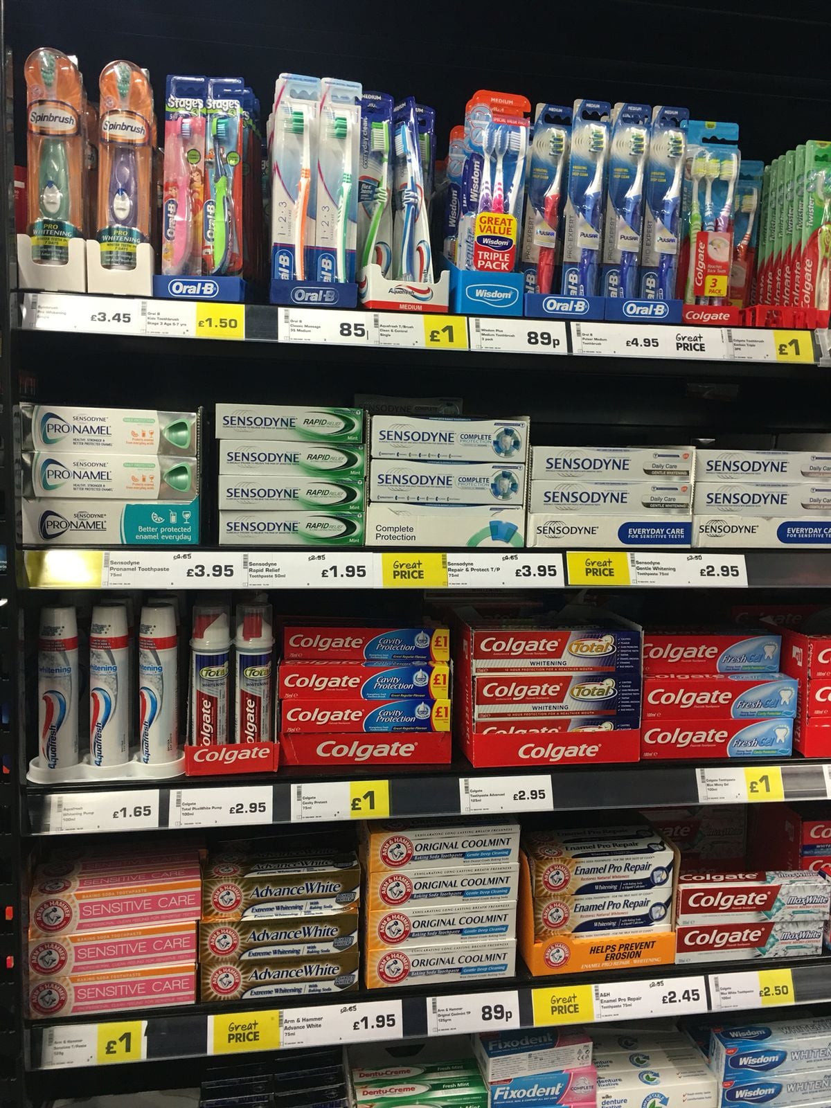 A supermarket shelf displaying various toothbrushes and toothpastes, including brands like Oral-B, Sensodyne, Colgate, and Aquafresh, with yellow price tags and clear product labeling.