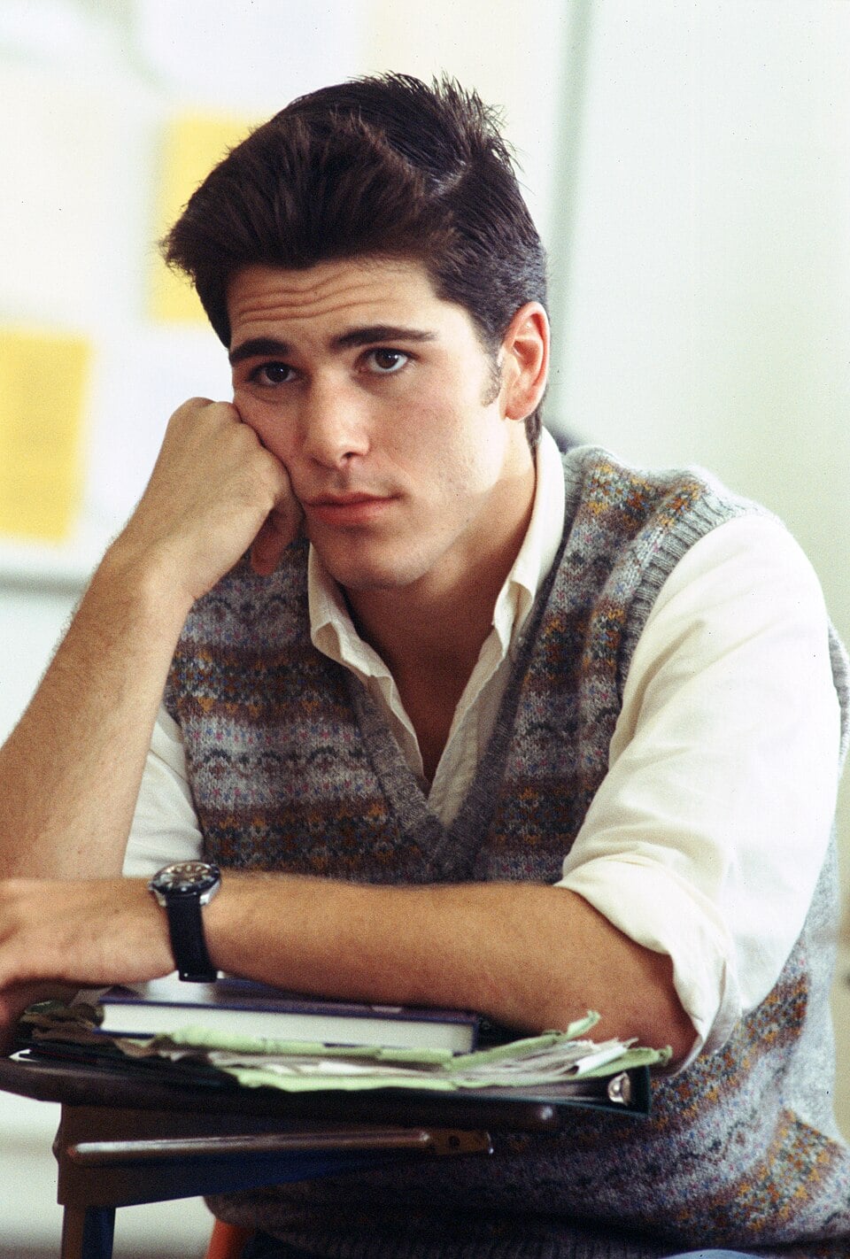 A young man with dark hair sits at a desk, resting his head on his hand. He wears a patterned sweater vest over a white shirt and looks thoughtfully at the camera. Papers and folders are on the desk in front of him.