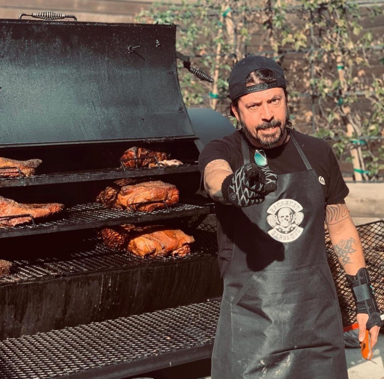 A man wearing a black cap, black shirt, and apron stands in front of a large outdoor smoker filled with smoked meat, pointing towards the camera with a gloved hand.
