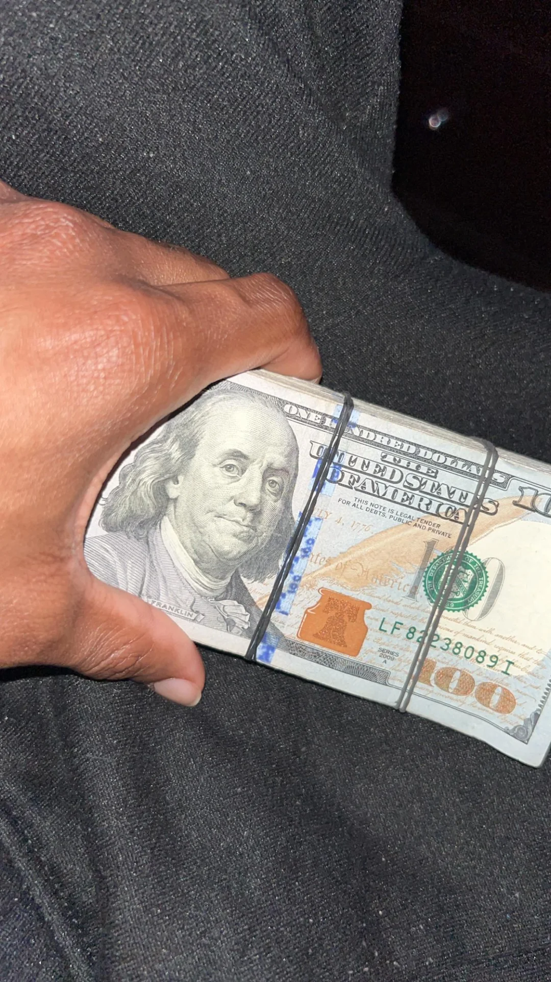 A hand holding a thick stack of U.S. one hundred dollar bills, secured with rubber bands, against a dark fabric background.