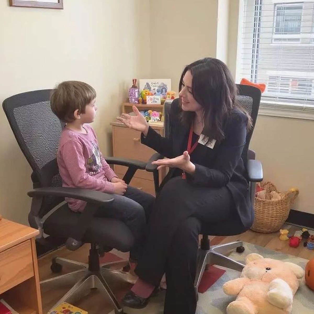 A woman in professional attire sits in an office chair, smiling and talking to a young boy seated next to her. The room has toys, books, and sunlight streaming in through the window.