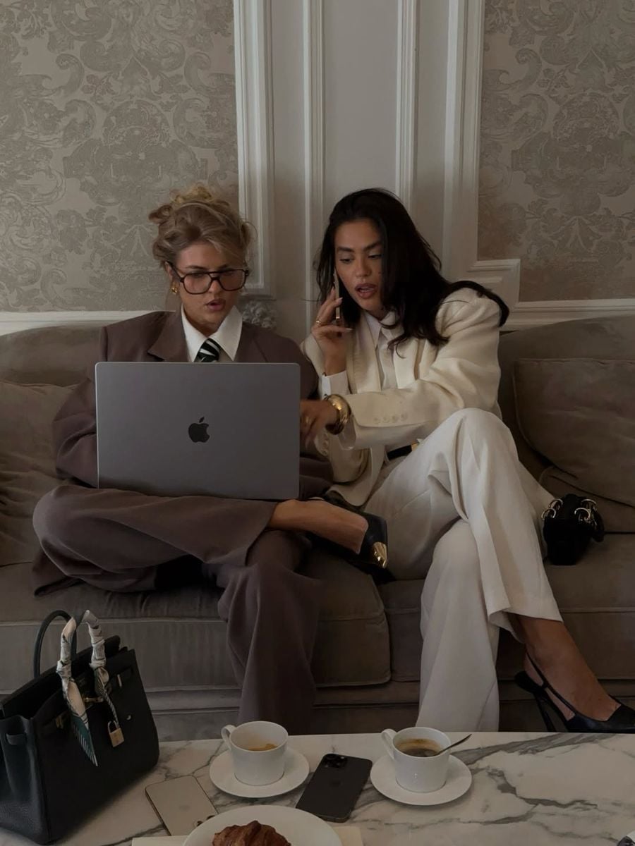 Two women in business attire sit on a sofa; one uses a laptop, the other talks on the phone. Both look focused. Coffee cups, a croissant, and handbags are on the table in front of them. The setting appears elegant and professional.