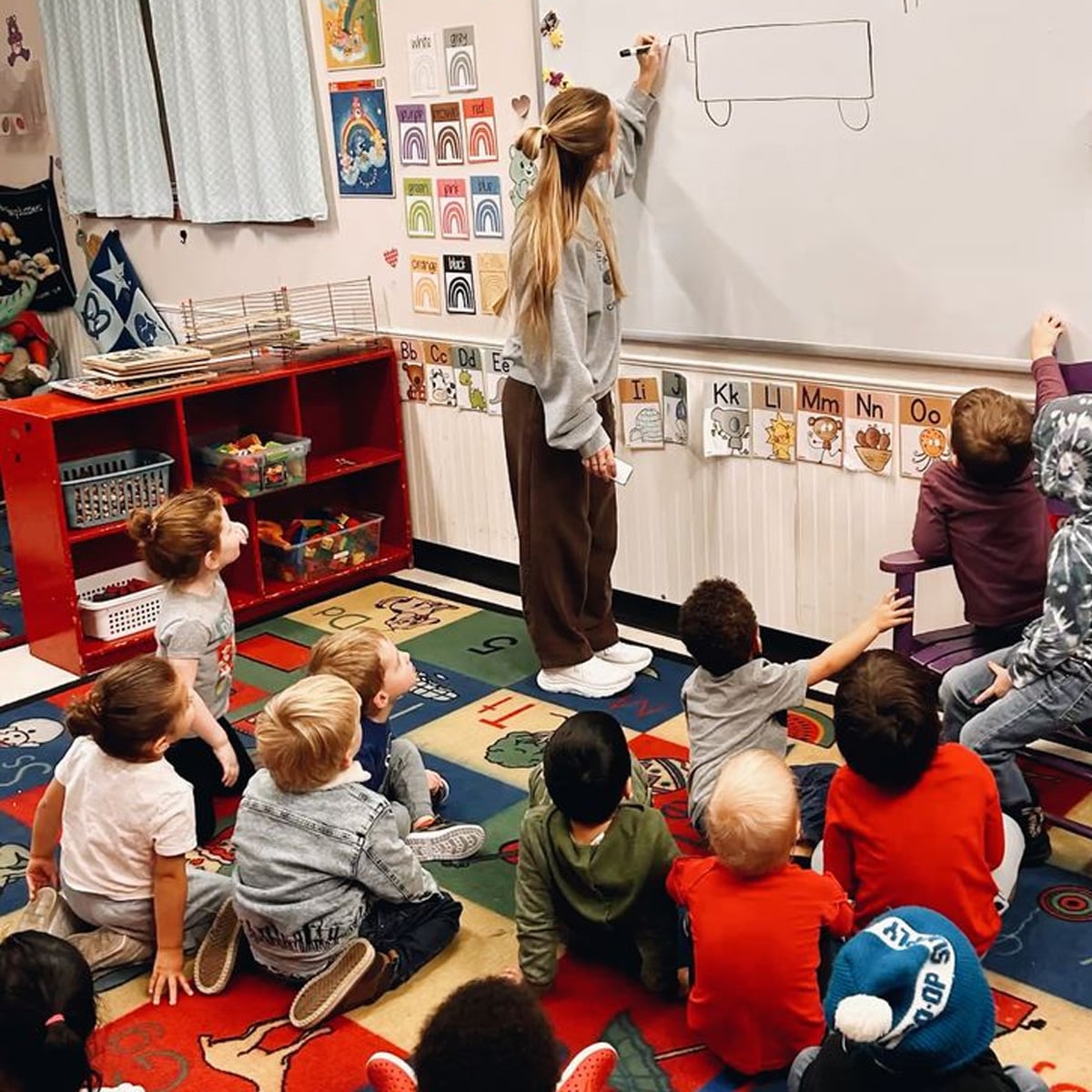 A teacher draws a bus on a whiteboard while a group of young children sit on a colorful classroom rug, watching and raising their hands to participate.