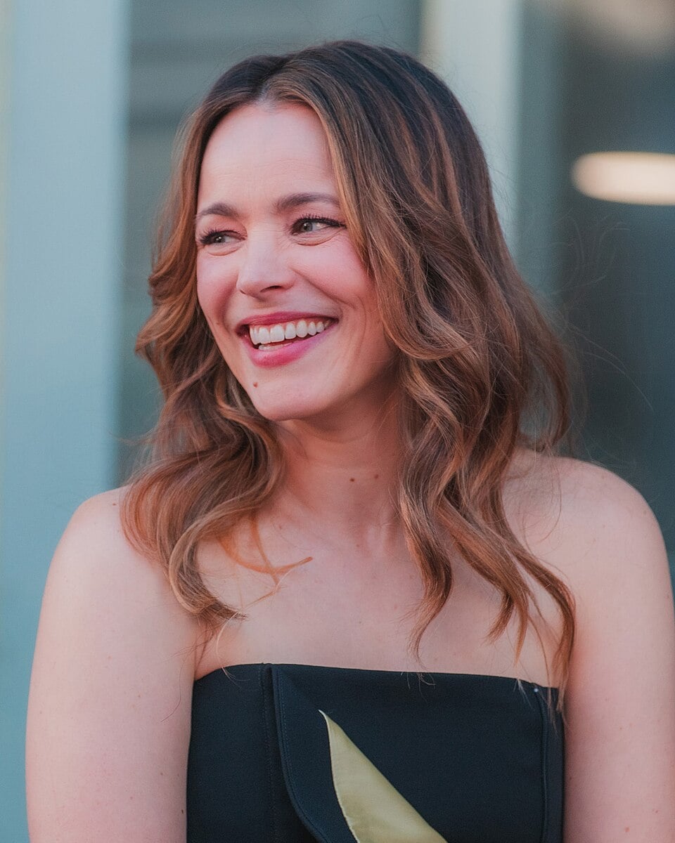 A woman with wavy, light brown hair smiles while looking to the side. She is wearing a strapless black top and is outdoors, with a blurred background of glass and light.