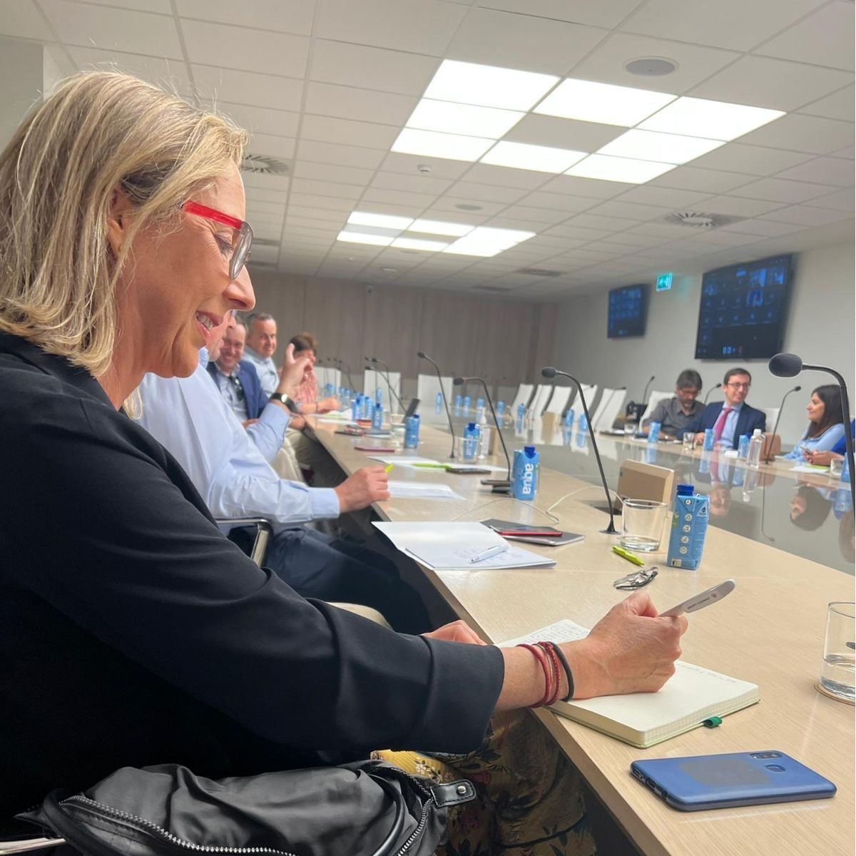 A woman with blonde hair and red glasses smiles while writing in a notebook at a long conference table filled with people, microphones, water bottles, and electronic devices in a modern meeting room.