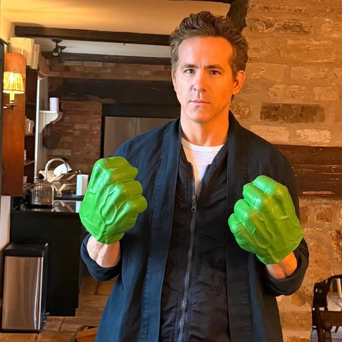 A man stands in a kitchen, wearing a dark jacket and white shirt, holding up his fists with oversized green Hulk gloves, looking directly at the camera with a neutral expression.